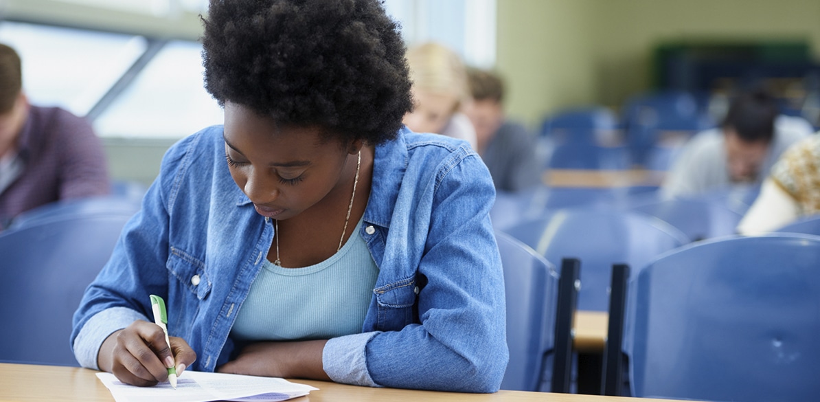 Educación Plus promueve el acceso al empleo de jóvenes de Costa Rica Joven estudiante concentrada escribiendo en un examen dentro de un aula, vestida con camisa vaquera azul y rodeada de otros alumnos al fondo.