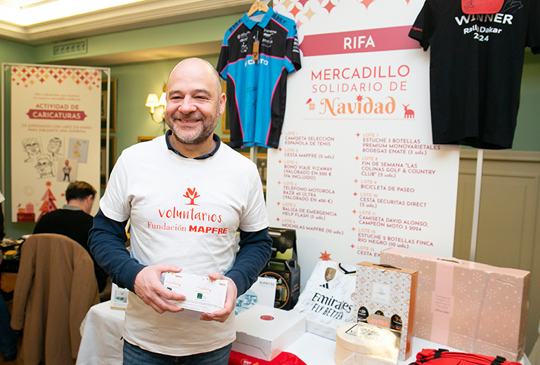 Voluntario sonriente de Fundación MAPFRE sosteniendo boletos de una rifa durante el Mercadillo Solidario de Navidad, con mesas, premios y carteles informativos del evento al fondo.