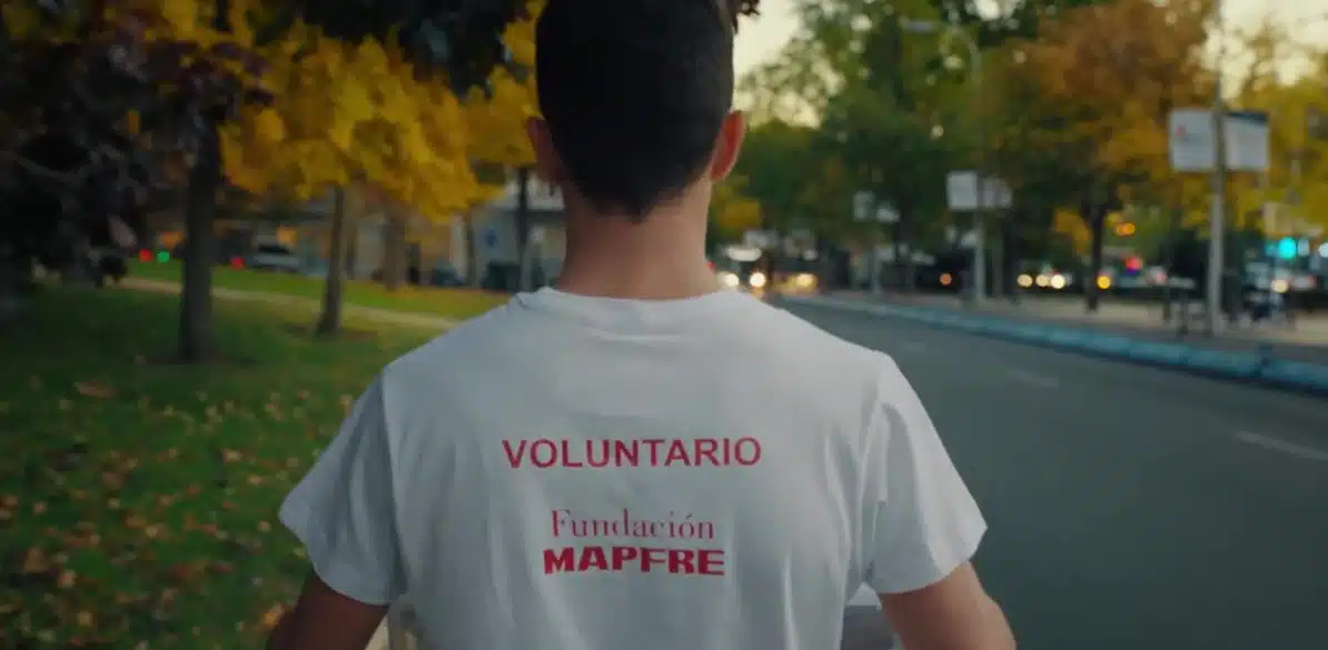 Volunteering A volunteer wearing a white t-shirt with 'VOLUNTARIO' and Fundación MAPFRE logo walks in an autumn park with yellow and green leaves, promoting acción social and community engagement.