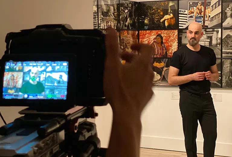 Arte en digital Un hombre calvo y con barba, vestido de negro, habla frente a una cámara en una galería de arte. La pantalla de la cámara muestra su imagen y parte de la exposición fotográfica de fondo con retratos y escenas urbanas coloridas.