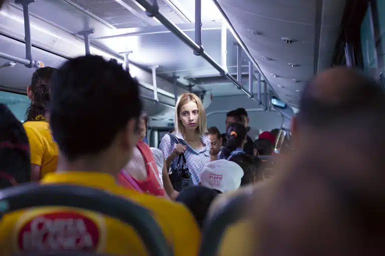 Interior de un autobús lleno de pasajeros, con una mujer rubia de pie en el centro mirando hacia la cámara entre asientos y personas desenfocadas.