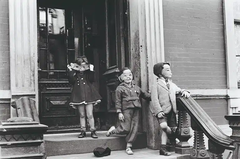 Fotografía en blanco y negro de Helen Levitt, c. 1940, con tres niños jugando con máscaras de papel en la entrada de un edificio de Nueva York; ellos están apoyados en la barandilla, y observan relajados la escena urbana.