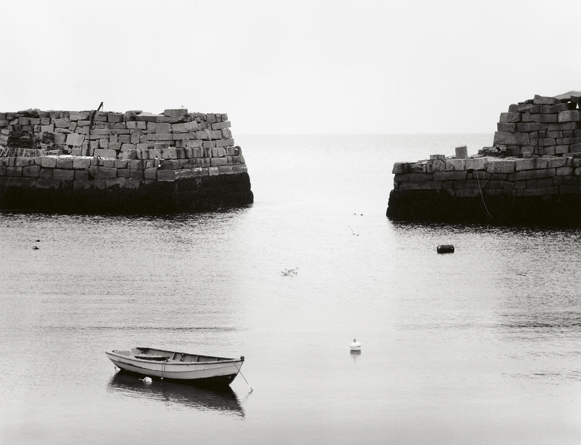 Boat on calm waters between two stone piers in black and white.