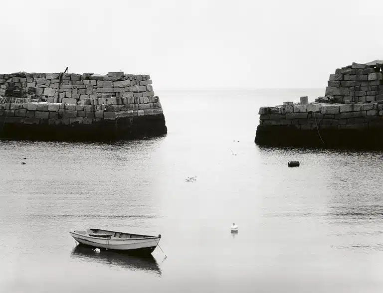 Minor White Boat on calm waters between two stone piers in black and white.