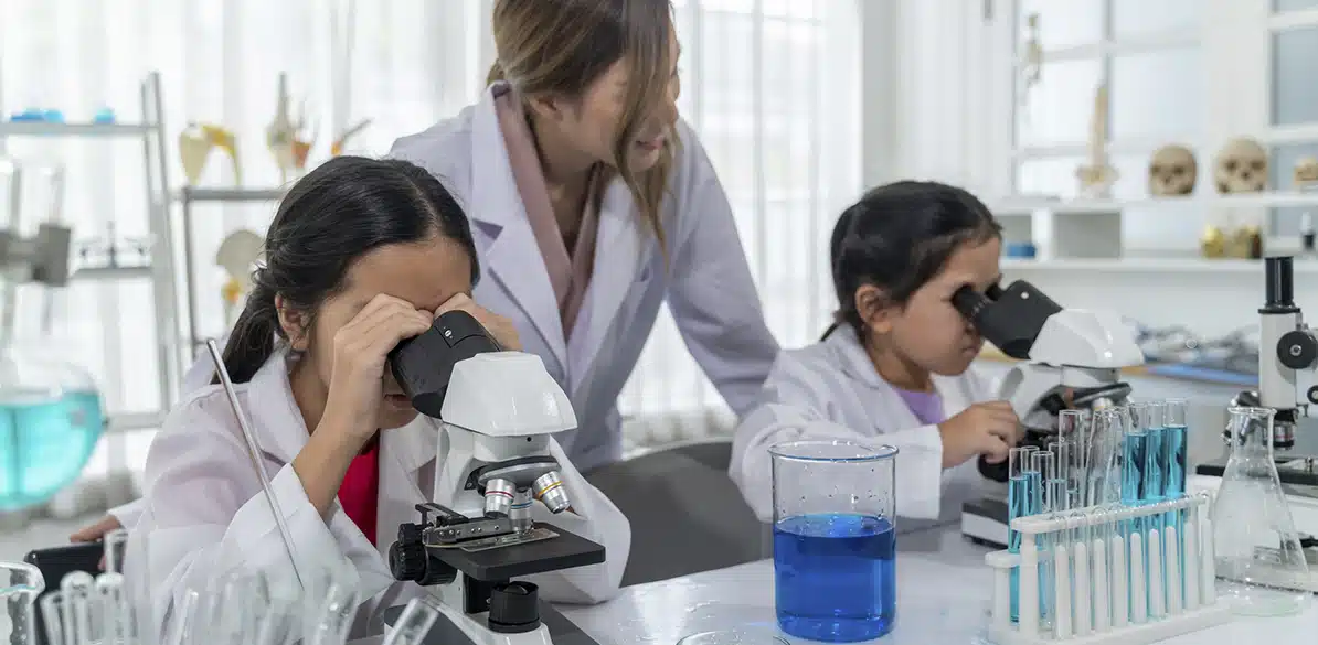Profesora supervisando a dos niñas con bata blanca mientras observan muestras en microscopios en un laboratorio escolar con tubos de ensayo y líquidos azules sobre la mesa.