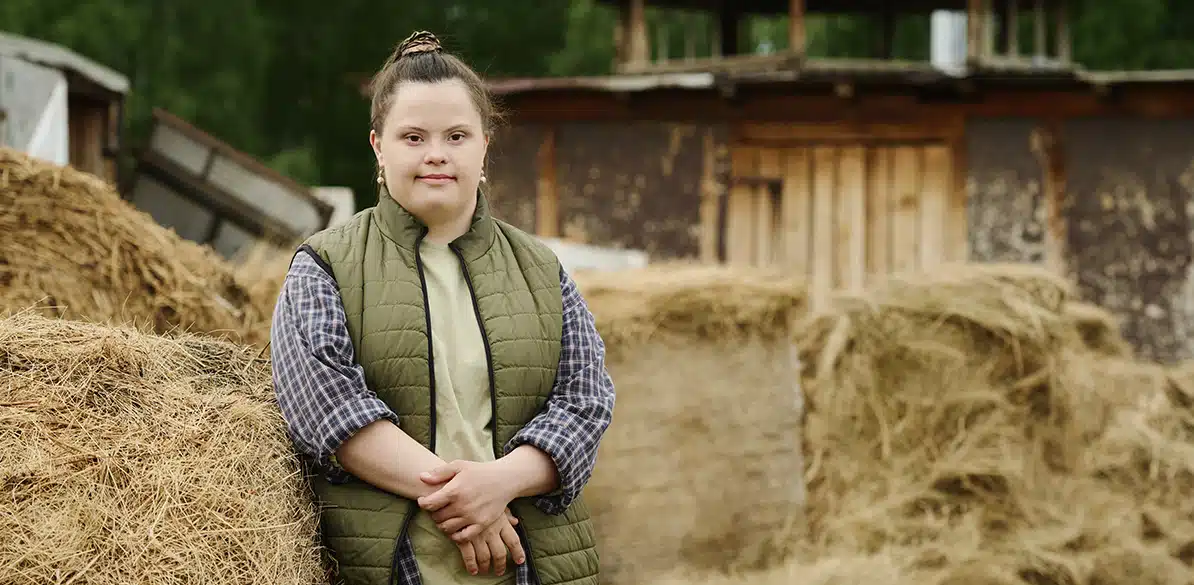 Joven con síndrome de Down posando con expresión serena junto a balas de heno en un entorno rural, con un chaleco acolchado verde y camisa de cuadros, con edificio de madera al fondo.