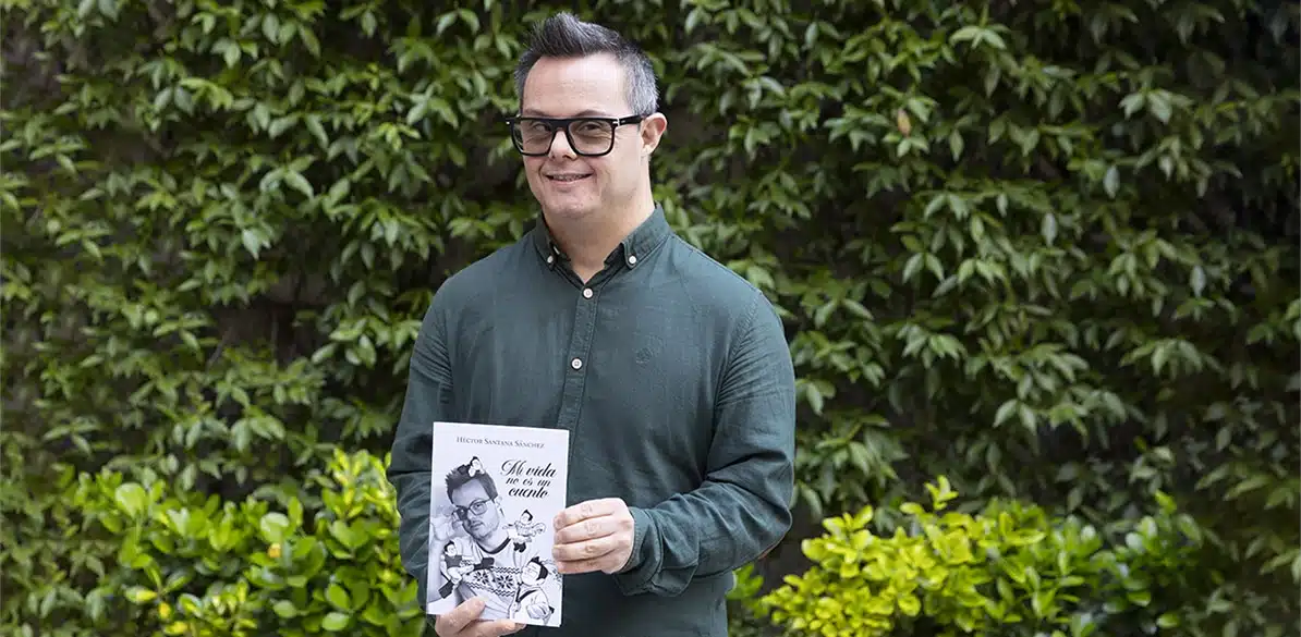 Héctor Santana Héctor Santana, un joven con síndrome de Down y gafas negras, sonriendo mientras sostiene su libro autobiográfico "Mi vida no es un cuento" frente a una pared de vegetación verde.