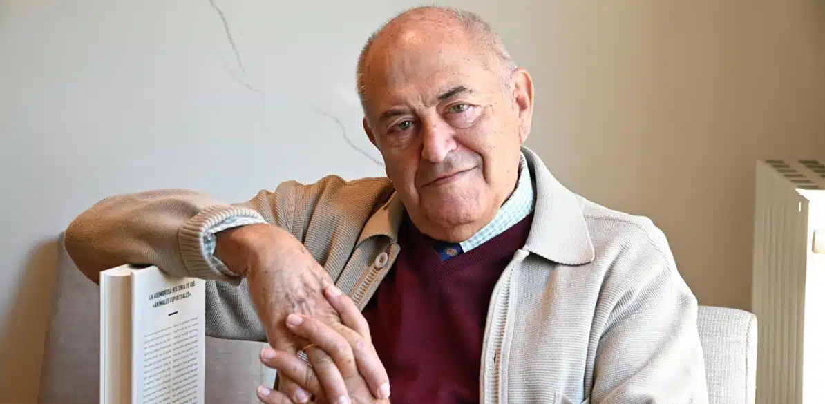 José Antonio Marina Retrato de un hombre mayor, de cabello canoso y piel arrugada, que luce una sonrisa suave mientras posa sentado. Lleva una camiseta roja, una camisa de cuello azul claro y un suéter beige, con un detalle de un libro abierto parcialmente visible a su lado derecho. Está mirando hacia la cámara con una expresión tranquila y relajada, mientras sus manos se entrelazan frente a él.