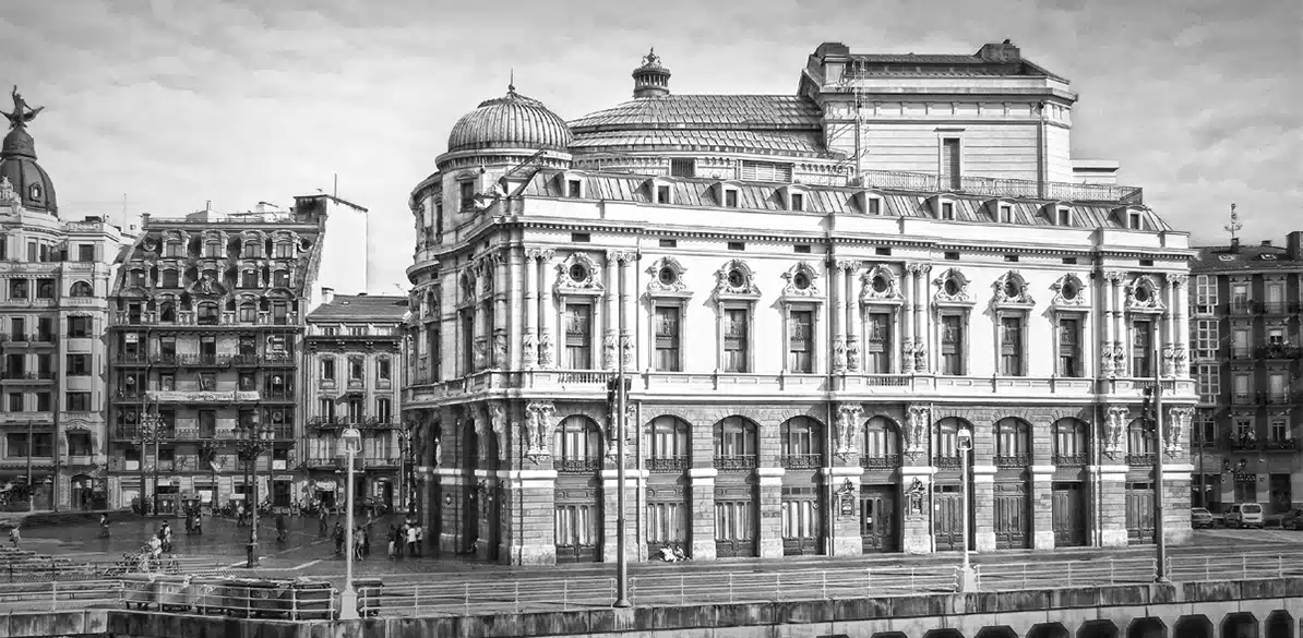 Imagen en blanco y negro del Teatro Arriaga en Bilbao, un edificio neobarroco con cúpulas y fachada ornamentada. A su alrededor se ven edificios antiguos y personas caminando junto a la ría, creando una escena urbana histórica y viva.