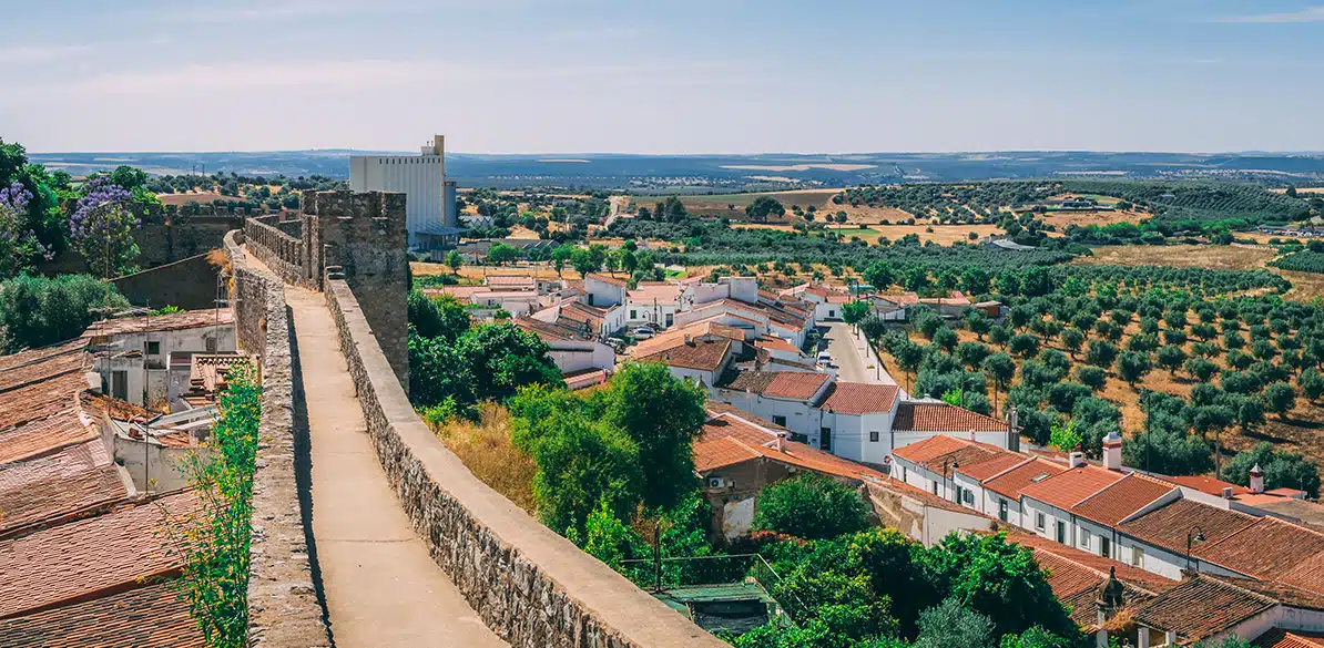 Red Ibérica Vista panorámica de un pueblo rodeado de olivares y campos bajo un cielo despejado, con murallas de piedra en primer plano y casas de tejados rojos al fondo, reflejando el compromiso del proyecto Red Ibérica con el apoyo a fundaciones españolas y portuguesas en entornos rurales.