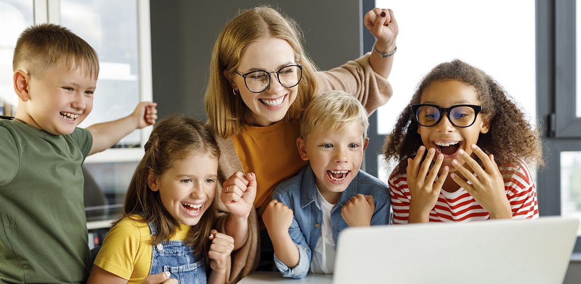 Profesora y grupo de niños celebrando juntos mientras están frente a un ordenador portátil.