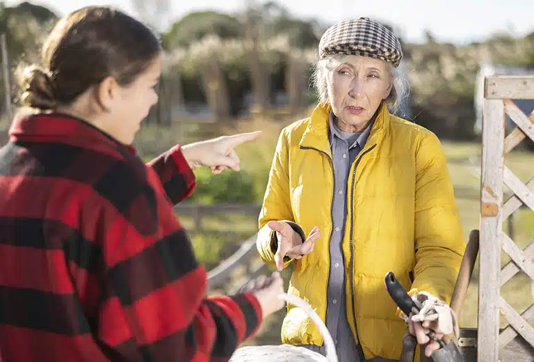 Alzheimer Andalucía Elderly woman wearing a yellow jacket and checkered cap in a rural setting, looking confused while a young woman next to her points and talks to her outdoors.