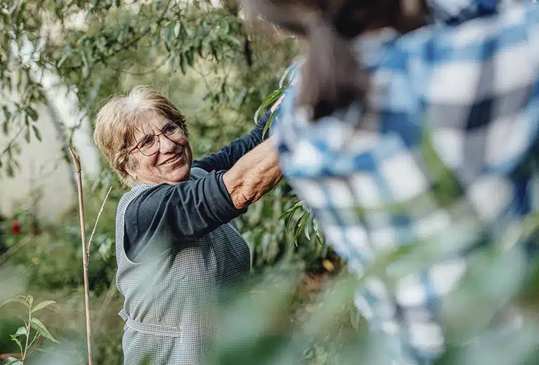 Alzheimer Ávila Smiling older woman wearing glasses and a long-sleeved shirt holds a tree branch while working in a vegetable garden, accompanied by another person.