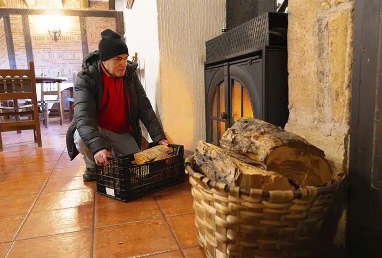 AMFORMAD Older man crouching next to a lit wood stove, placing a box of logs; wicker basket with firewood in the foreground in a rural house with a terracotta floor.