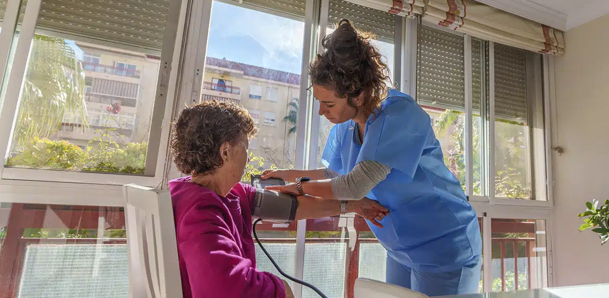 ASPAYM Madrid Healthcare professional in blue uniform takes the blood pressure of an elderly woman sitting at home, with natural light coming in through large windows.