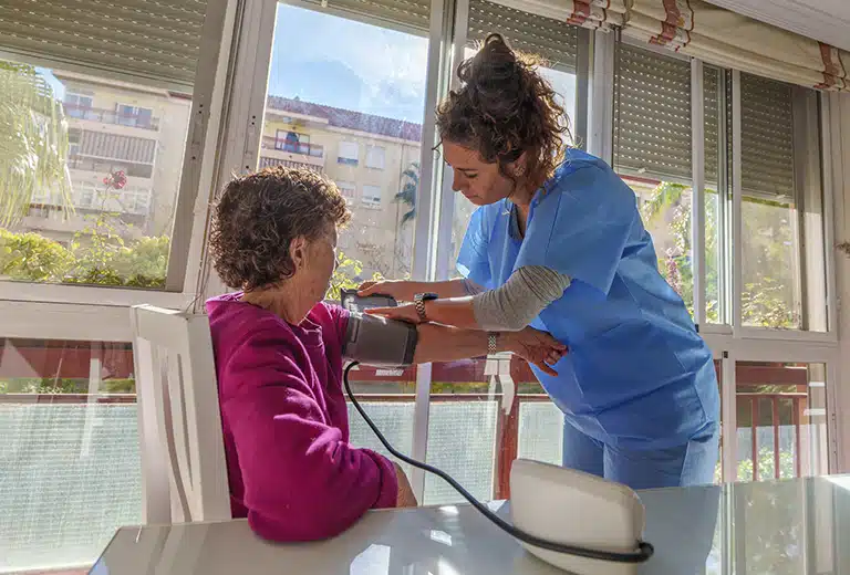 ASPAYM Madrid Healthcare professional in blue uniform takes the blood pressure of an elderly woman sitting at home, with natural light coming in through large windows.