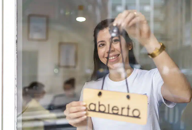 Cáritas Diocesana Mérida/Badajoz Woman smiling as she hangs an “open” sign on the door of her store in a rural area.