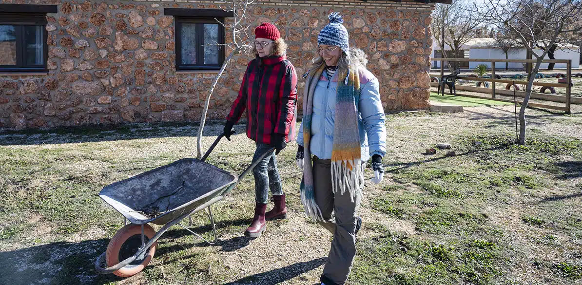 COCEDER Two women walk through a rural field; one is pushing a wheelbarrow while the other walk with her. Both are wearing warm clothing and are surrounded by nature.