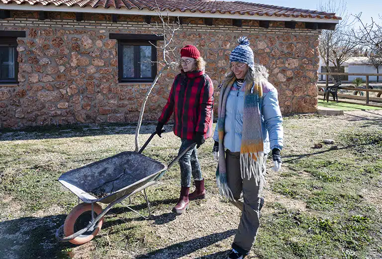 COCEDER Two women walk through a rural field; one is pushing a wheelbarrow while the other walk with her. Both are wearing warm clothing and are surrounded by nature.