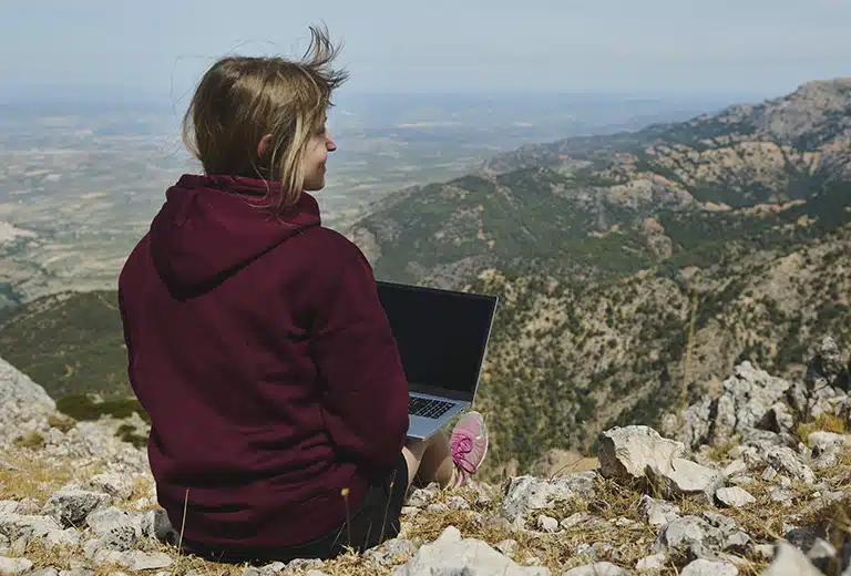 FUNDACIÓN DKV INTEGRALIA A young woman working on a laptop at the top of a mountain, with a panoramic view of mountains and valleys around her.