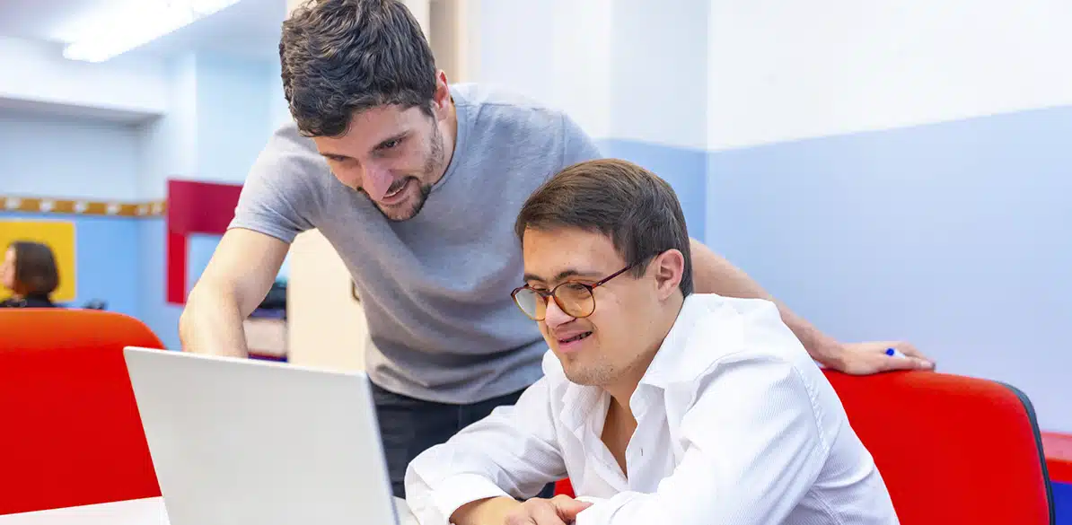 DOWN ESPAÑA Person with Down syndrome receiving help from a colleague to work on a computer in a modern office environment