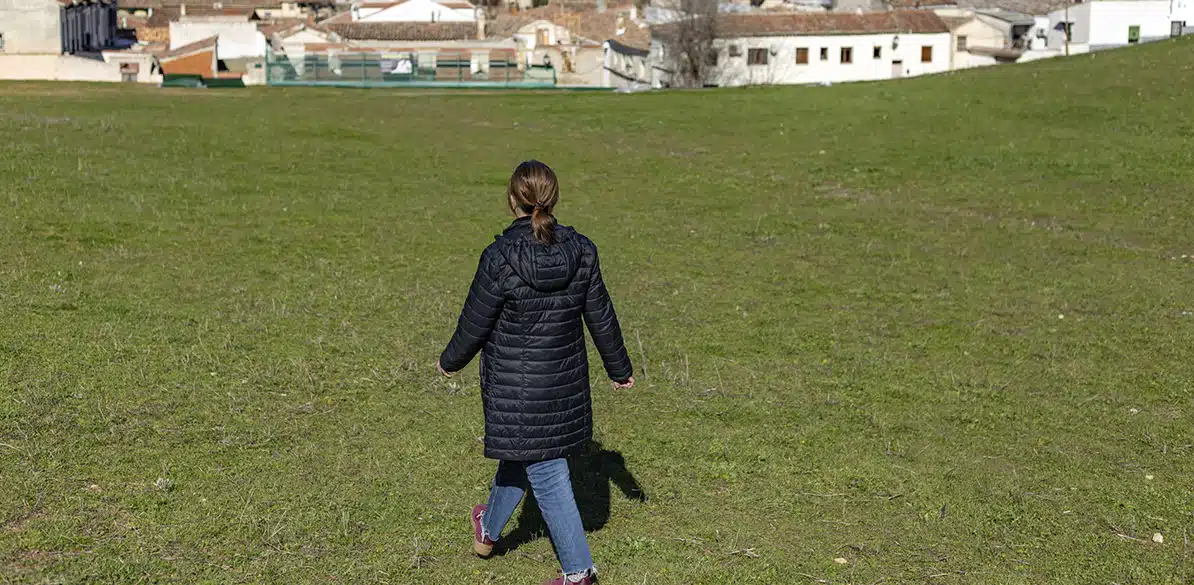 Fundación ECCA Social Young woman walking alone through a green field in a rural area, with houses in the distance. She is wearing a black coat and sneakers while enjoying the landscape.