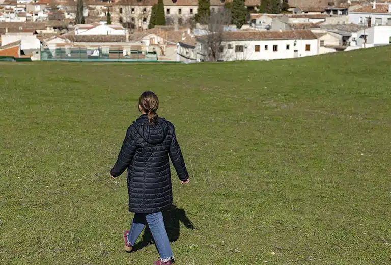 Fundación ECCA Social Young woman walking alone through a green field in a rural area, with houses in the distance. She is wearing a black coat and sneakers while enjoying the landscape.