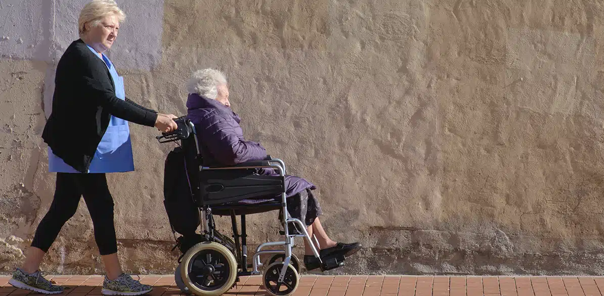 El Saliente Association Healthcare professional pushes an elderly woman in a wheelchair along a street next to a wall, symbolizing local care and support for dependency.