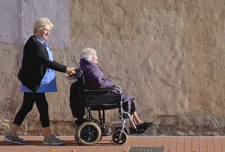 Asociación El Saliente Healthcare professional pushes an elderly woman in a wheelchair along a street next to a wall, symbolizing local care and support for dependency.