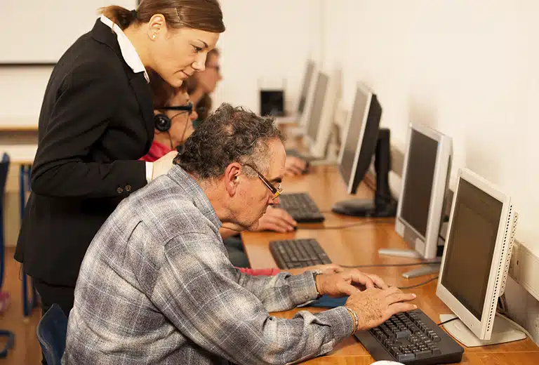 Fundación Purísima Concepción An older person wearing glasses receives help while using a computer in a classroom, assisted by a professional who is providing digital guidance and training.