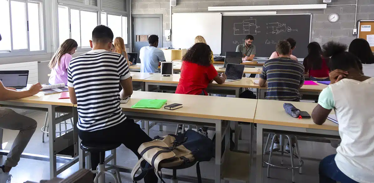 INCISO Classroom with students sitting at high tables using laptops and taking notes; teacher at the front next to a blackboard with diagrams, natural light coming through windows.