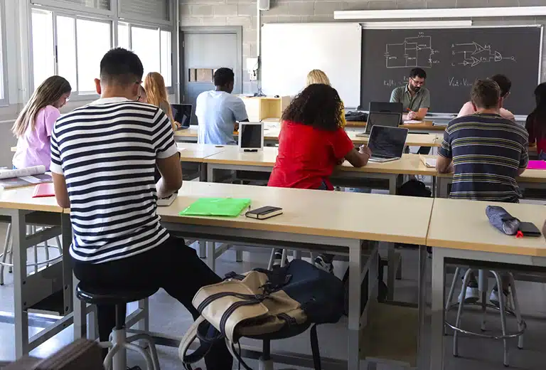 INCISO Classroom with students sitting at high tables using laptops and taking notes; teacher at the front next to a blackboard with diagrams, natural light coming through windows.