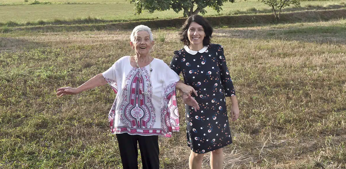 Fundación Intras, Arcadia, Isol Smiling older woman in a white printed blouse walks through a field of dry grass while a younger woman in a dark dress takes her arm in a rural setting.