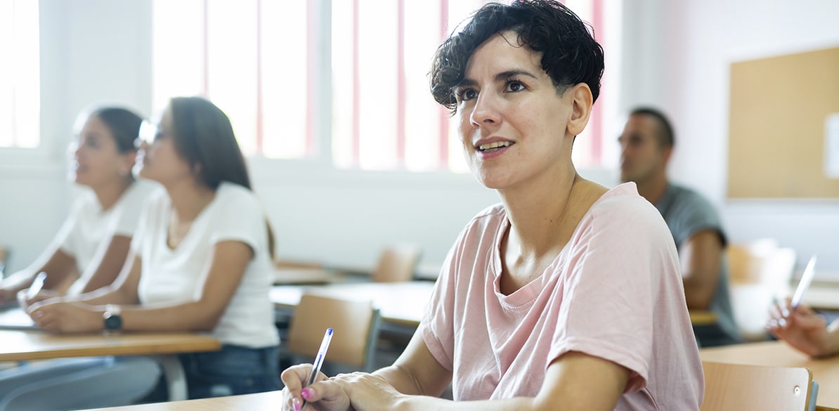Fundación San Cebrian Woman with short hair sitting in a classroom, taking notes with a pen while attending a training session; other people out of focus in the background.