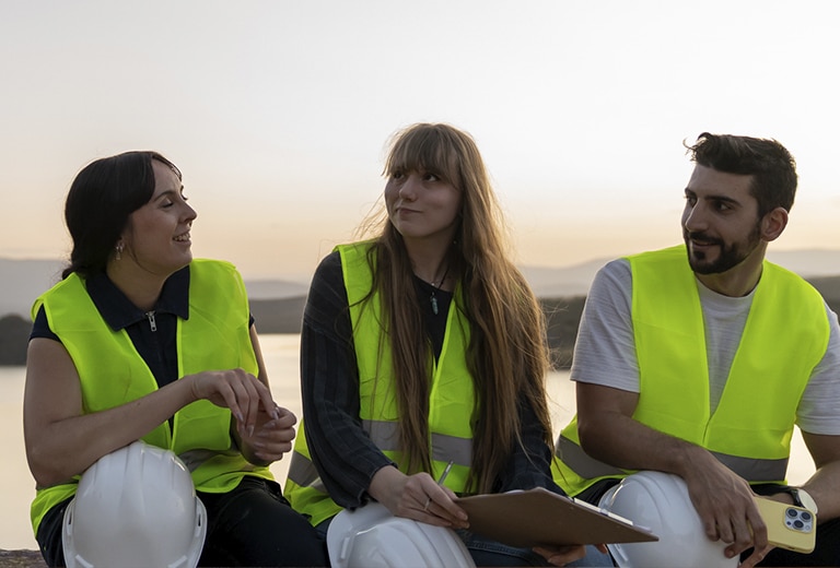 Santa María La Real Three people sitting outdoors, wearing reflective safety vests and helmets, chat while enjoying the natural landscape, symbolizing teamwork in rural areas.