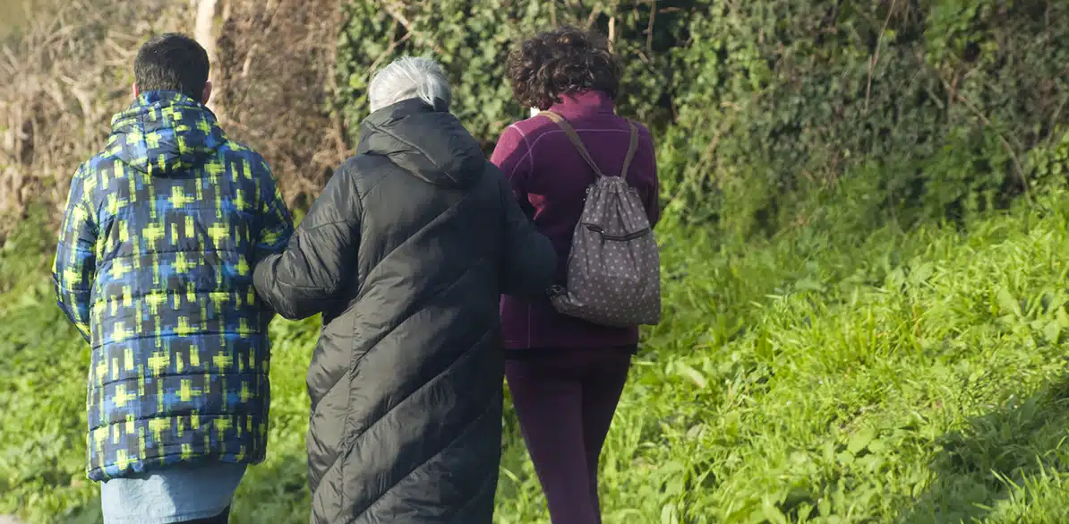 Santa María La Real Three people walk along a rural path next to some greenery with their backs to the camera: an older person wearing a black coat is accompanied by two people, one wearing a blue patterned jacket and the other carrying a backpack.