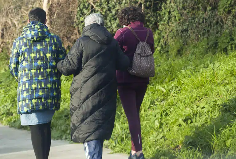 Santa María La Real Three people walk along a rural path next to some greenery with their backs to the camera: an older person wearing a black coat is accompanied by two people, one wearing a blue patterned jacket and the other carrying a backpack.