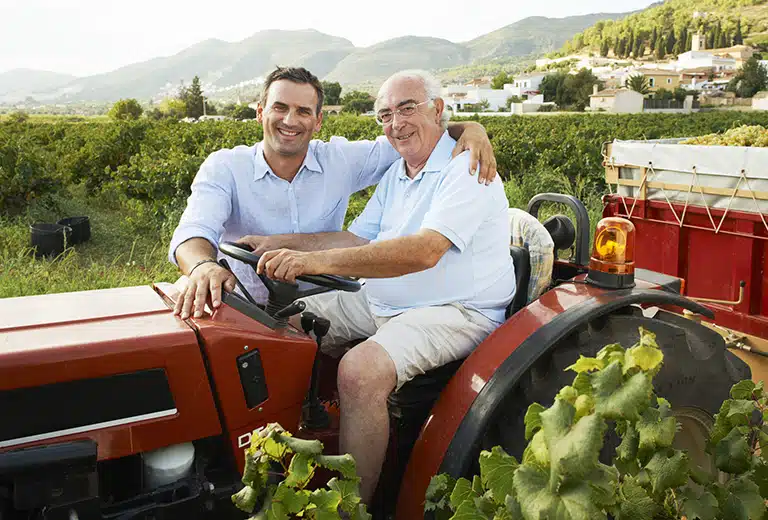 Two men smile as they sit on a red tractor in a vineyard, with hills and farmhouses in the background. It reflects agricultural work, closeness, and life in the rural environment.