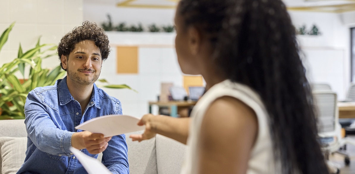Dos personas en una oficina moderna, con una de ellas, un hombre joven sonriente de camisa azul, entregando papeles a una joven de pelo largo y blusa blanca.  Este podría ser momento clave en el proceso de solicitud a la ayuda del programa Taller de Aprendices 2025, de Fundación MAPFRE.