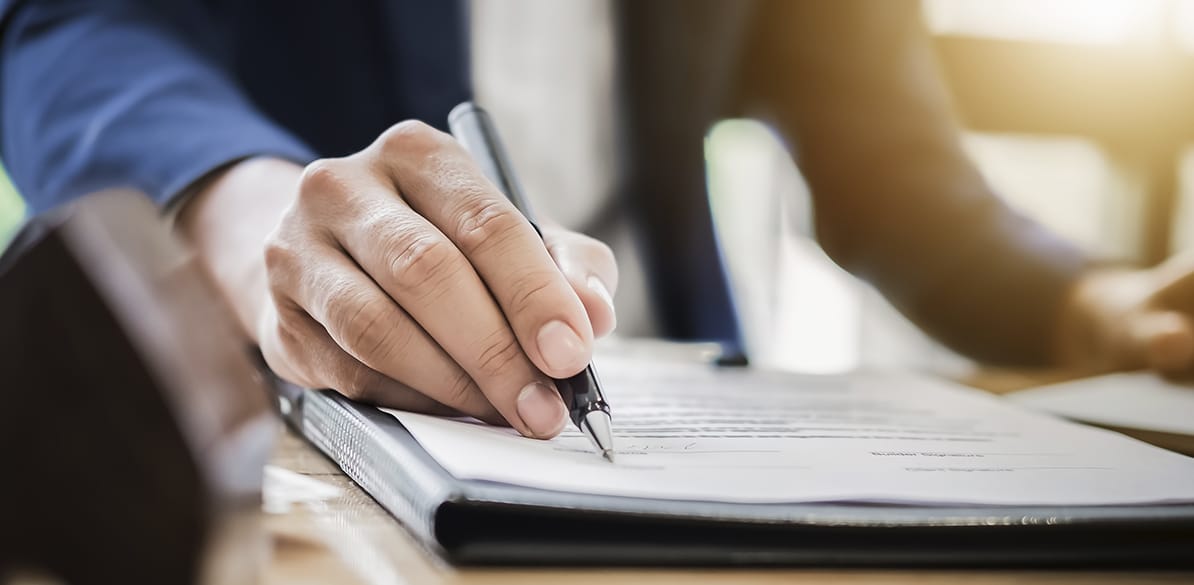 Mano firmando un documento con un bolígrafo negro, sobre una carpeta de piel. 