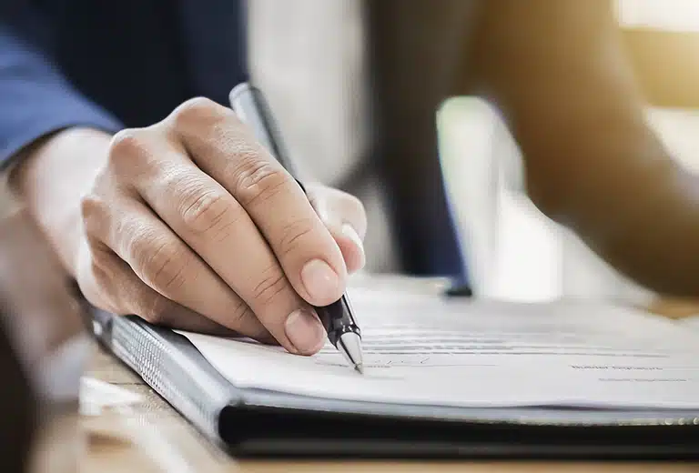 Mano firmando un documento con un bolígrafo negro, sobre una carpeta de piel. 
