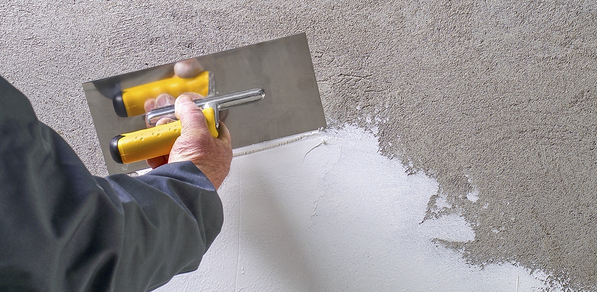 Bases de la convocatoria Persona aplicando yeso en una pared gris con una paleta de metal con mango amarillo. Uno de los trabajos de construcción y albañilería que se benefician de las ayudas para la contratación de aprendices para formación en oficios especializados.