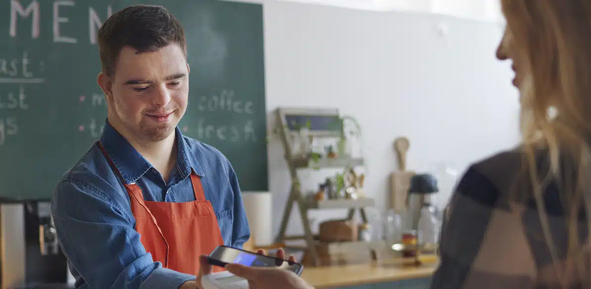 Hombre joven con discapacidad intelectual trabajando como camarero en una cafetería y realizando un cobro con datáfono a una clienta, en un entorno laboral inclusivo orientado a la inserción laboral.