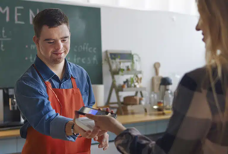 Hombre joven con discapacidad intelectual trabajando como camarero en una cafetería y realizando un cobro con datáfono a una clienta, en un entorno laboral inclusivo orientado a la inserción laboral.