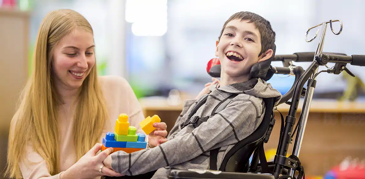 Enfermedades raras Niño con discapacidad sentado en silla de ruedas utilizando bloques de construcción durante una sesión de terapia asistida con una profesional, en un entorno educativo o de rehabilitación para mejorar su desarrollo y autonomía.