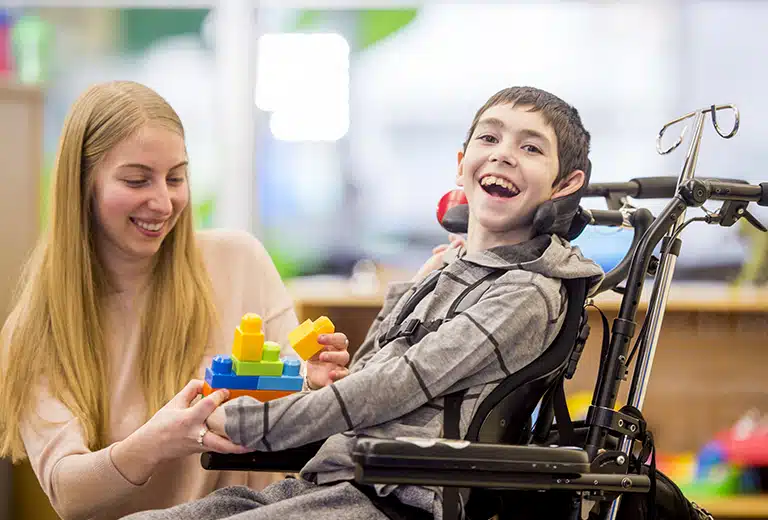 Niño con discapacidad sentado en silla de ruedas utilizando bloques de construcción durante una sesión de terapia asistida con una profesional, en un entorno educativo o de rehabilitación para mejorar su desarrollo y autonomía.