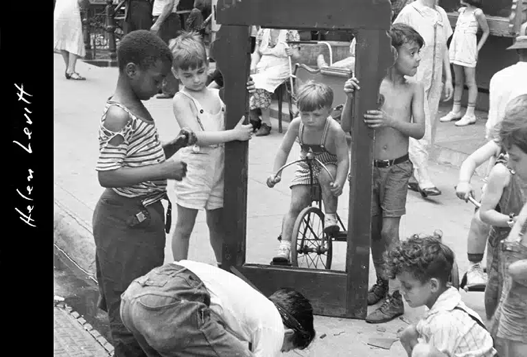 Helen Levitt Fotografía en blanco y negro de niños jugando en la calle, con un niño sosteniendo un marco de madera mientras otro pasa en bicicleta. La escena muestra una actividad infantil en un entorno urbano.