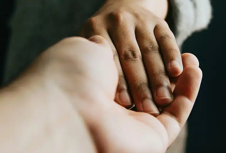 Salud mental Primer plano de dos manos entrelazadas: una más clara, que parece ofrecer apoyo, y otra más oscura, que recibe el gesto con suavidad. El fondo está desenfocado, y la imagen transmite el valor del acompañamiento y el apoyo interpersonal. Representa la importancia de crear vínculos seguros y de confianza como parte esencial de la salud mental en el aula.