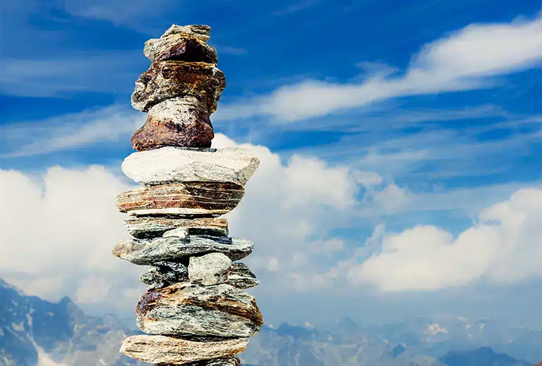 Fotografía de una torre de piedras cuidadosamente apiladas una sobre otra, en equilibrio vertical, con un cielo azul brillante y nubes al fondo. Las piedras varían en tamaño, forma y color. Al fondo se aprecian picos montañosos desenfocados, lo que refuerza la sensación de altura y solidez. La imagen representa visualmente el análisis comparativo que implica un ranking de reaseguradores, donde se evalúa la solidez y desempeño de cada entidad en un entorno global competitivo.