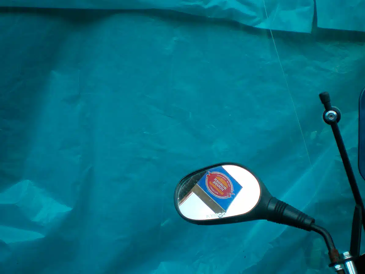 Abril Coudougnan Retrovisor de motocicleta refleja un cartel de comida rápida sobre un fondo azul turquesa arrugado.